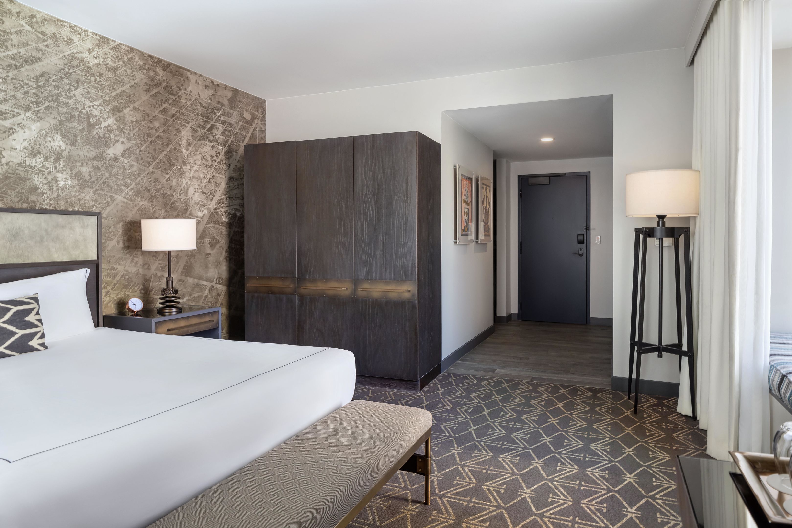 A hotel room with a bed, bench, nightstand, lamp, wardrobe, and patterned floor.