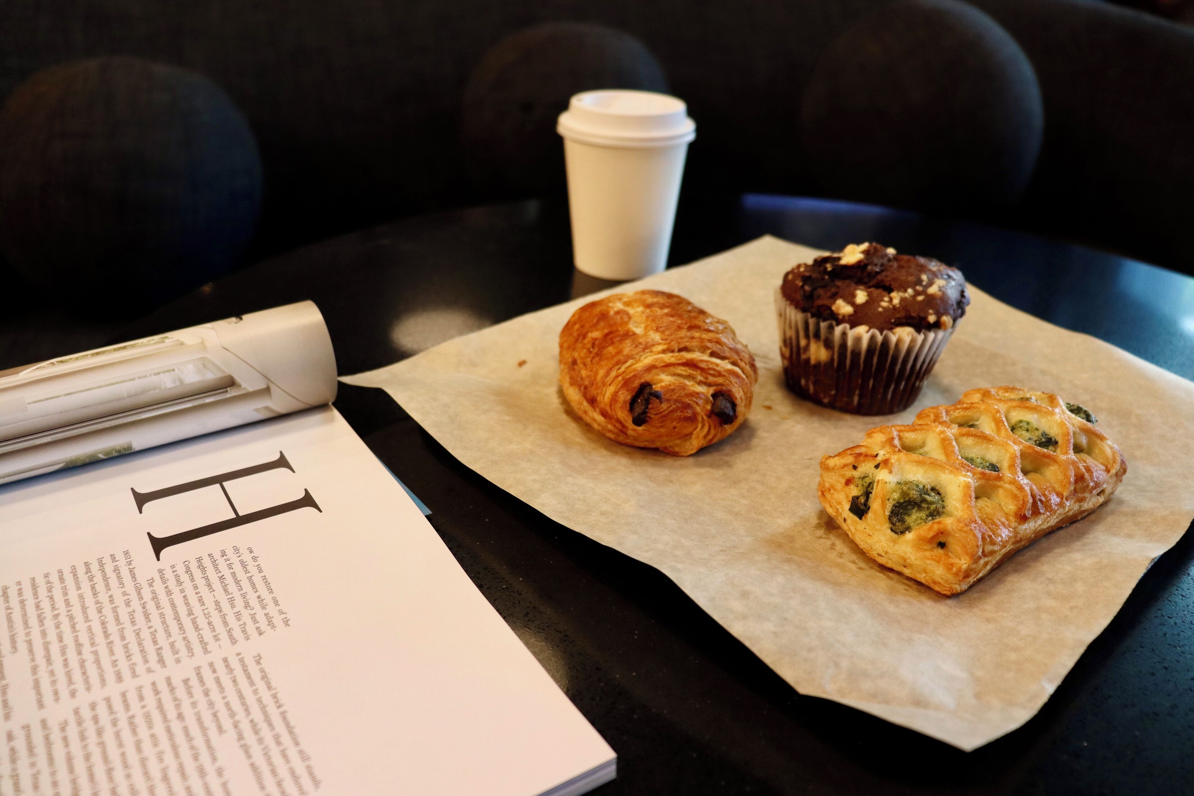 Una mesa con un croissant, un muffin y una taza de café, junto con un libro y un bolígrafo.