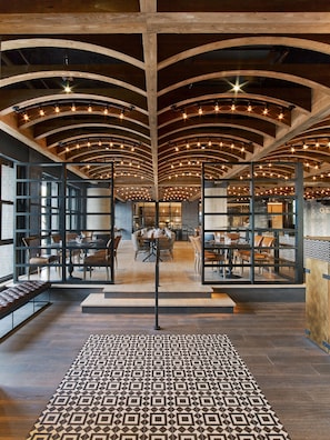 A spacious interior with high ceilings, wooden beams, and a patterned floor. Tables and chairs are arranged for dining.