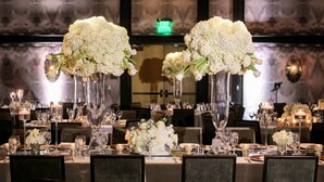 Un salón de banquetes con mesas dispuestas para un evento formal, destacando altos jarrones con flores blancas y centros de mesa con velas.