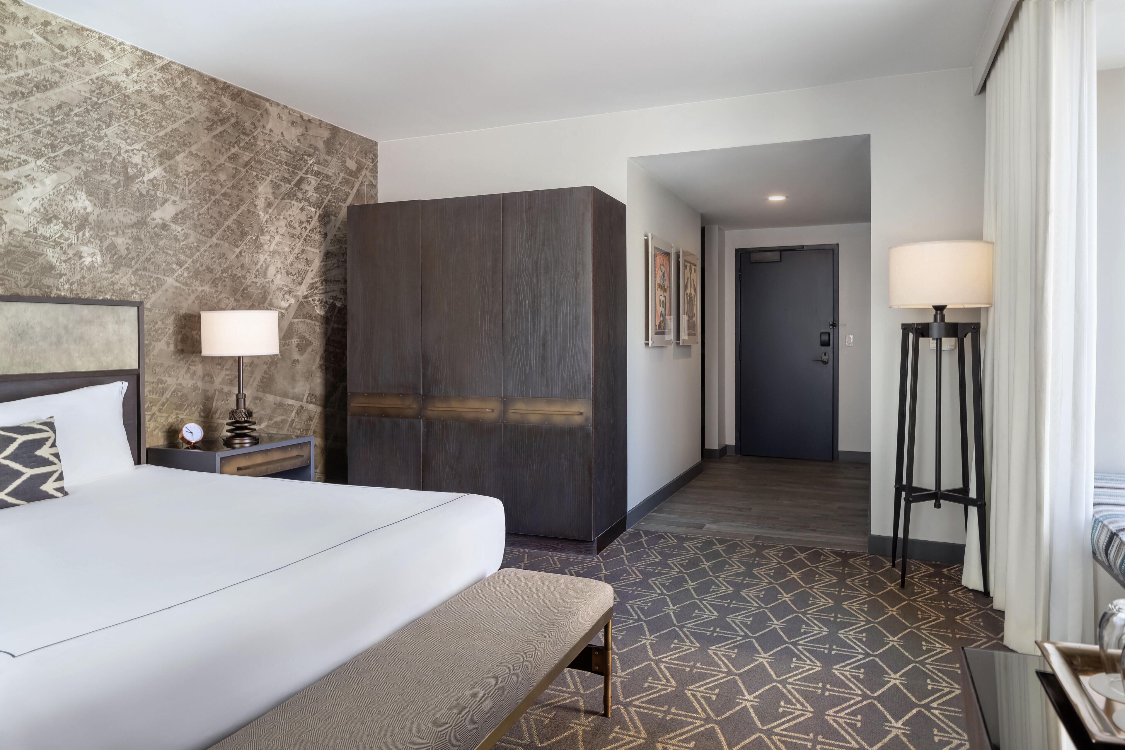 A hotel room with a large bed, a bench, a nightstand, a lamp, a wardrobe, and a patterned floor.