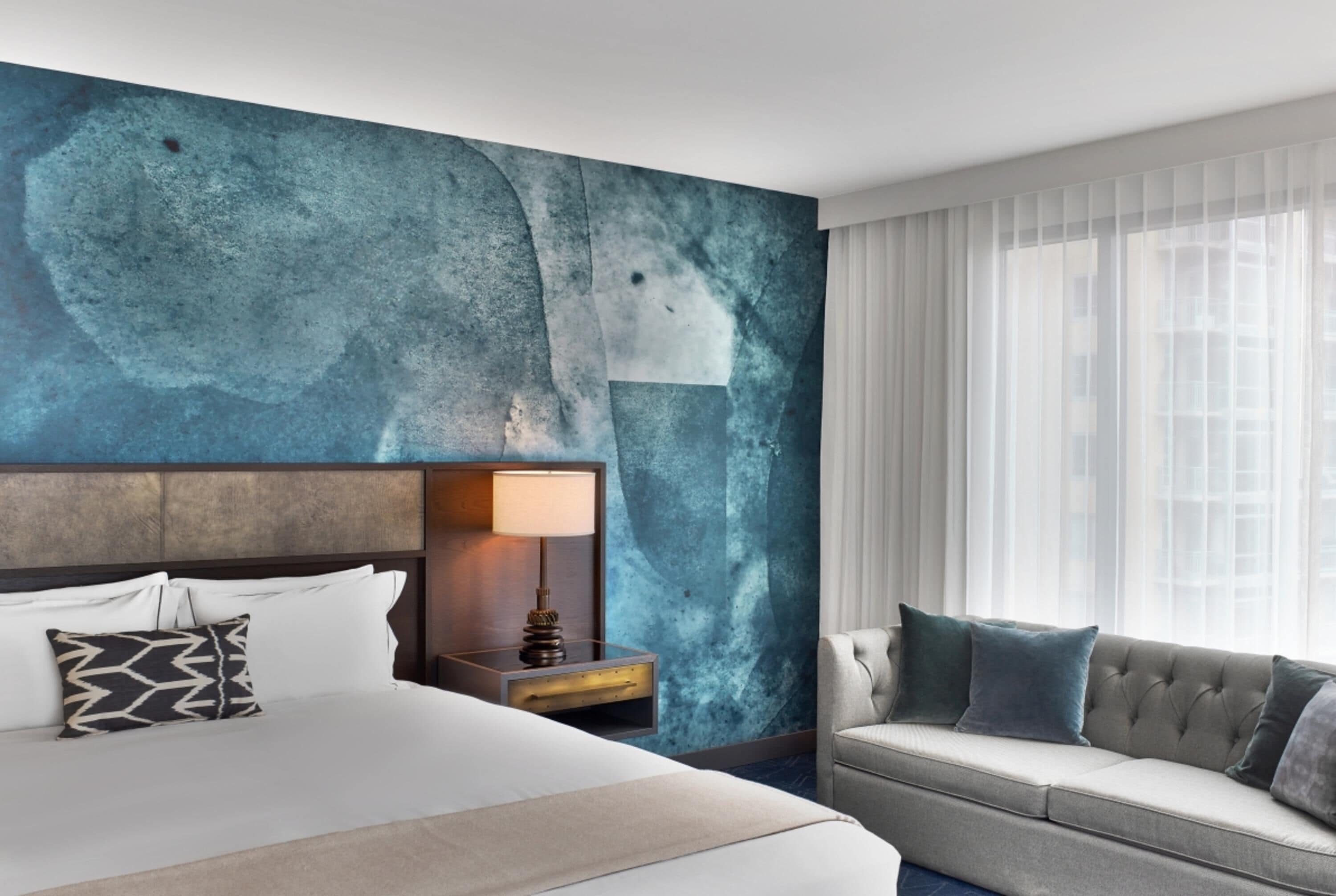A hotel room with a large bed, a sofa, a nightstand with a lamp, and a wall with a textured design.