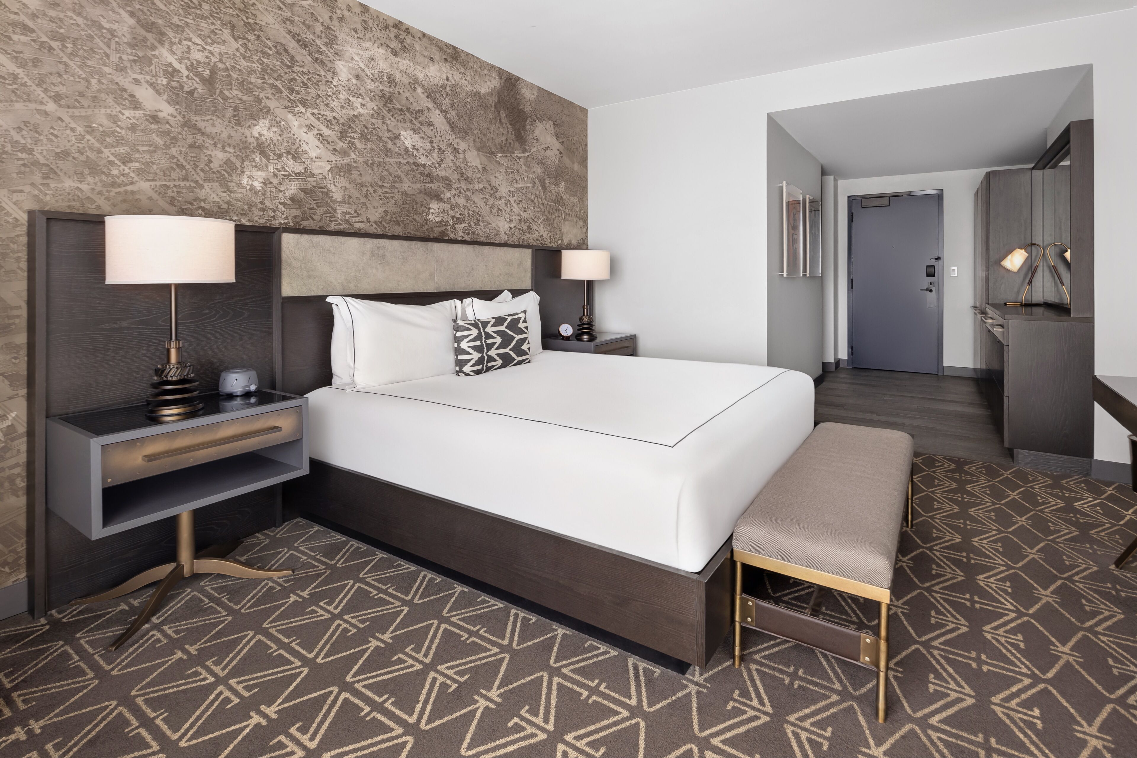 Habitación de hotel con una cama grande, mesitas de noche, un banco, un armario y una puerta que da a otra habitación.