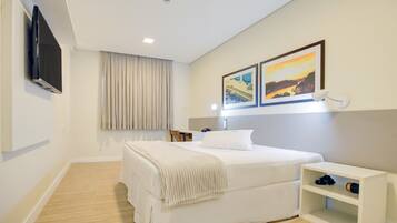 Una habitación de hotel con una cama, un televisor montado en la pared, una mesita de noche y una estantería con objetos encima.