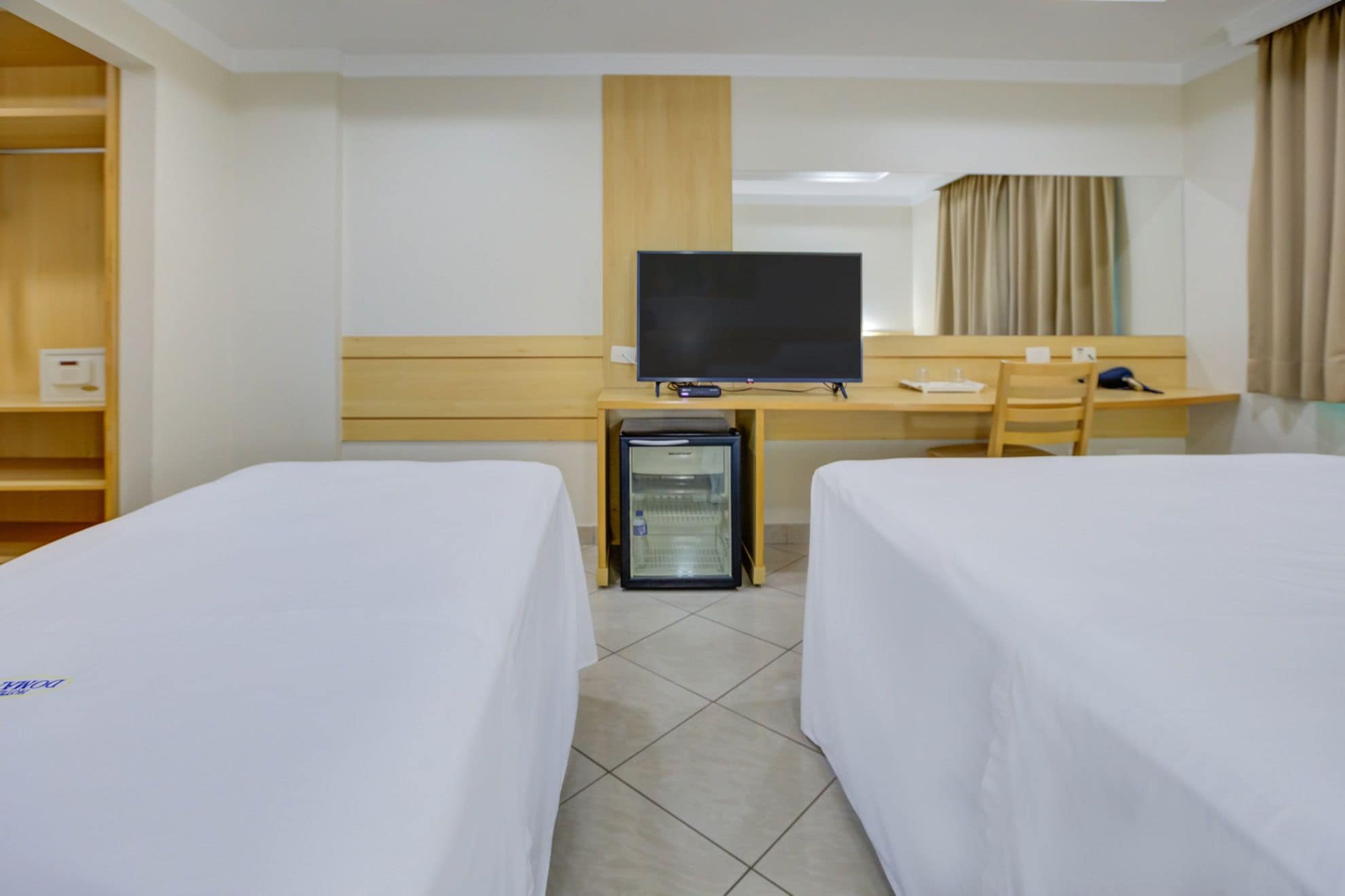A hotel room with two beds, a desk with a television, a mini-fridge, and a chair.