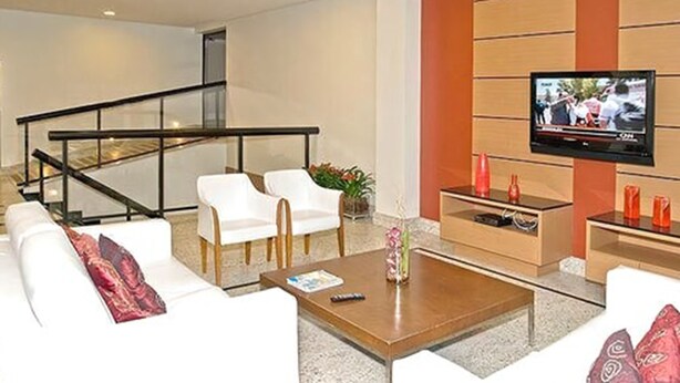 A modern living room with a sofa, chairs, a coffee table, and a TV mounted on the wall. - Hotel Domani (Guarulhos)