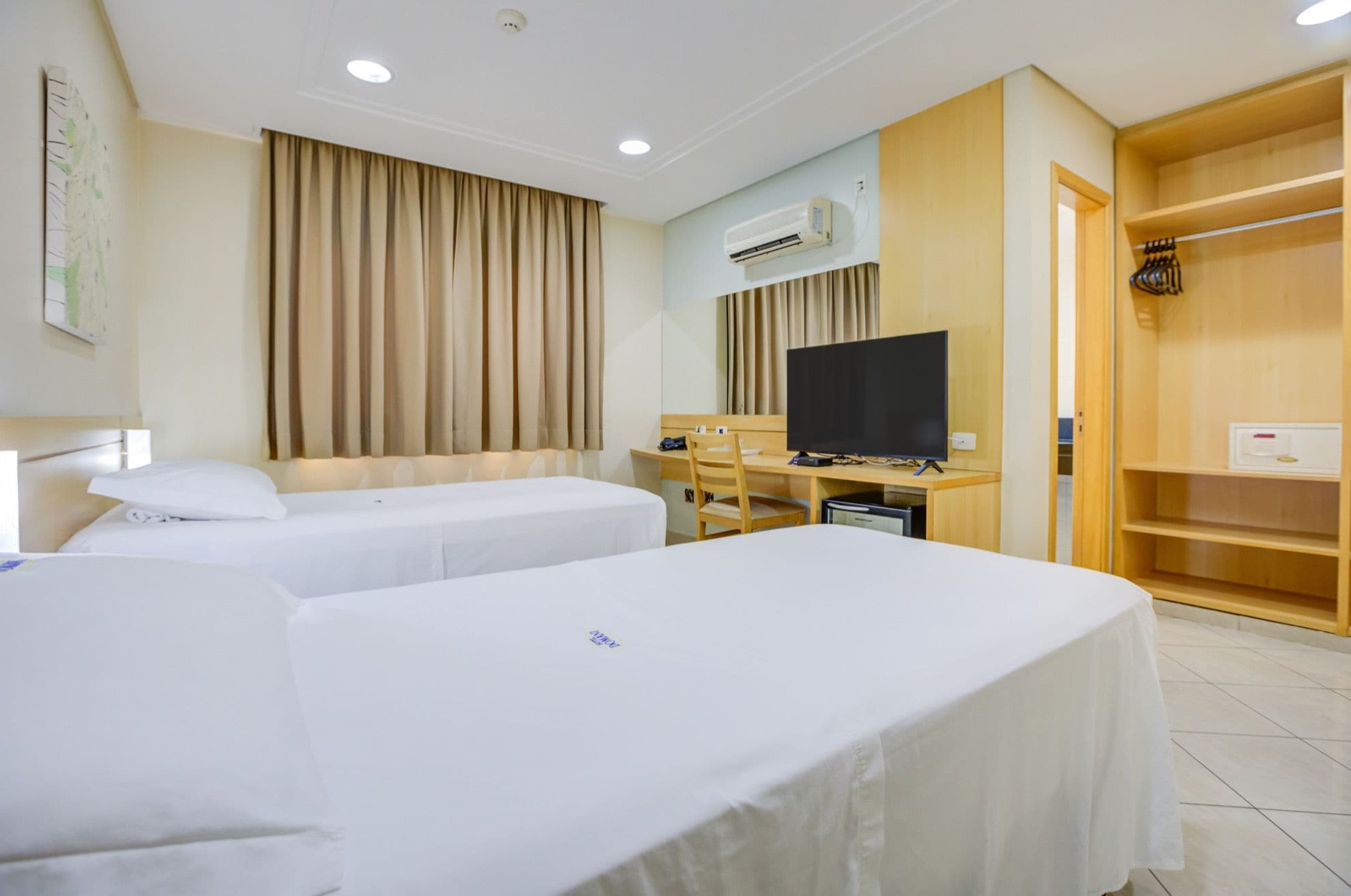A hotel room with two beds, a desk with a television, an air conditioning unit, and a wooden wardrobe.