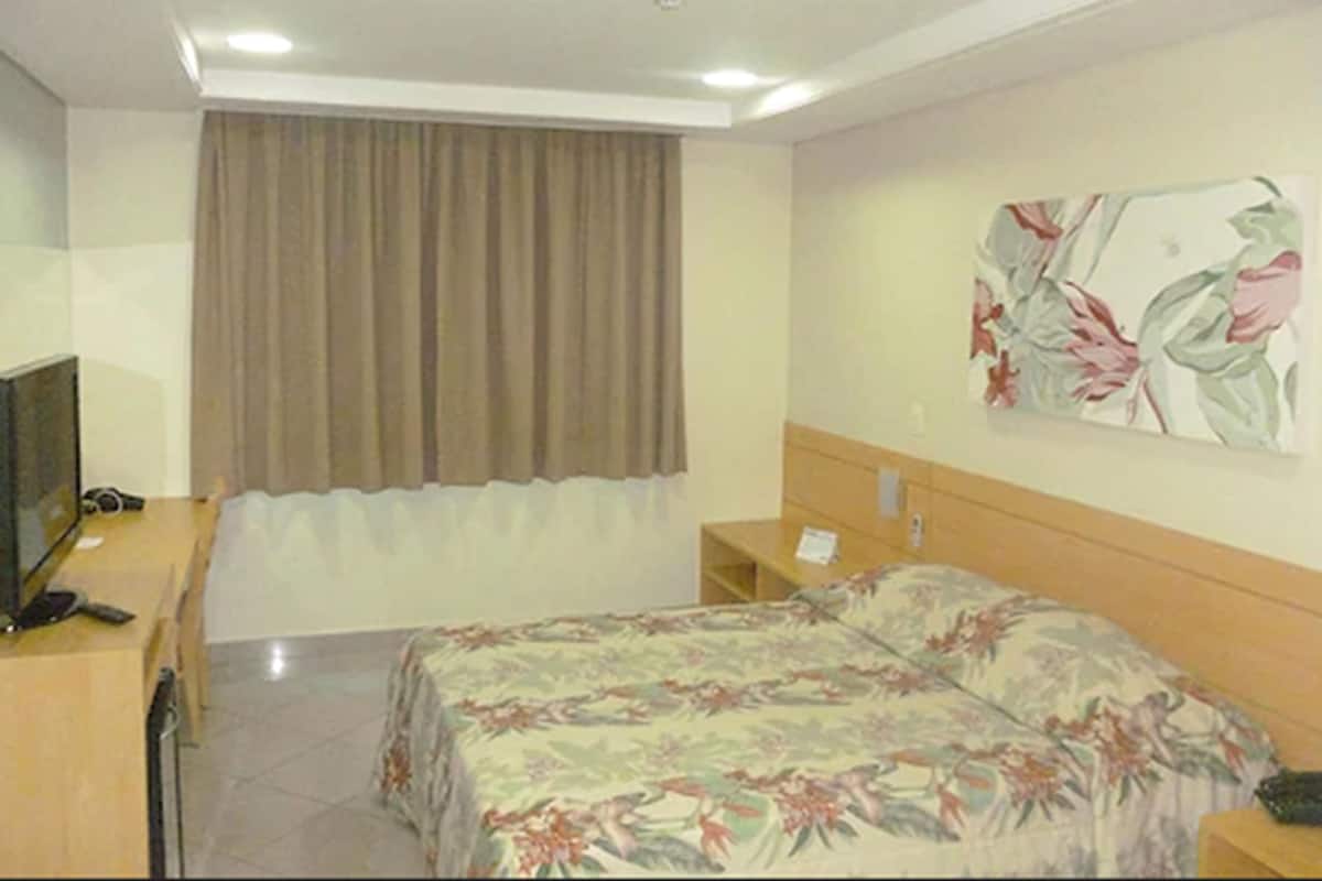 A hotel room with a bed, a desk, a chair, a television, a curtain, and a floral painting on the wall.