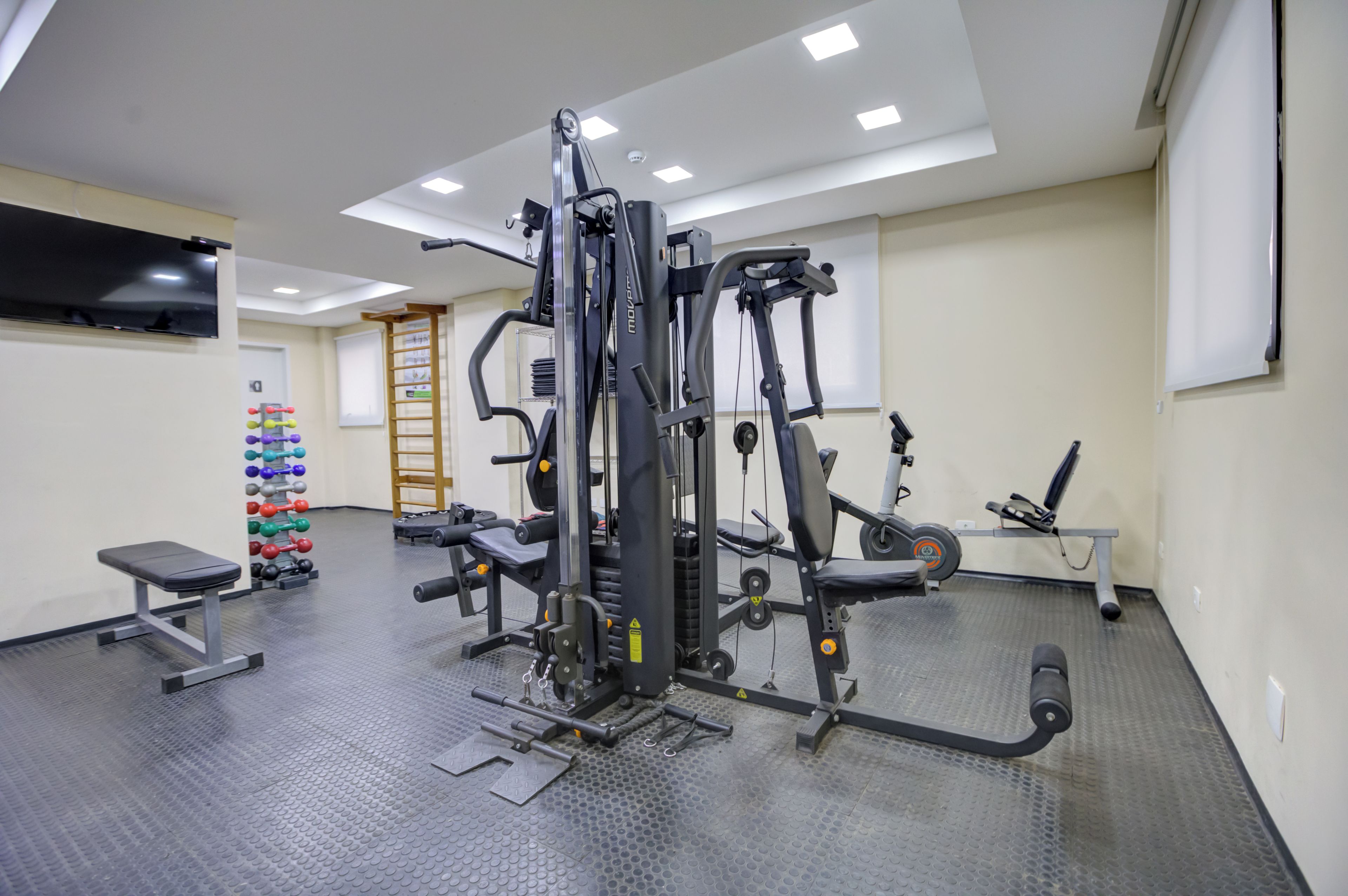 Uma academia com diversos equipamentos de exercício, uma TV fixada na parede e um sapateiro.