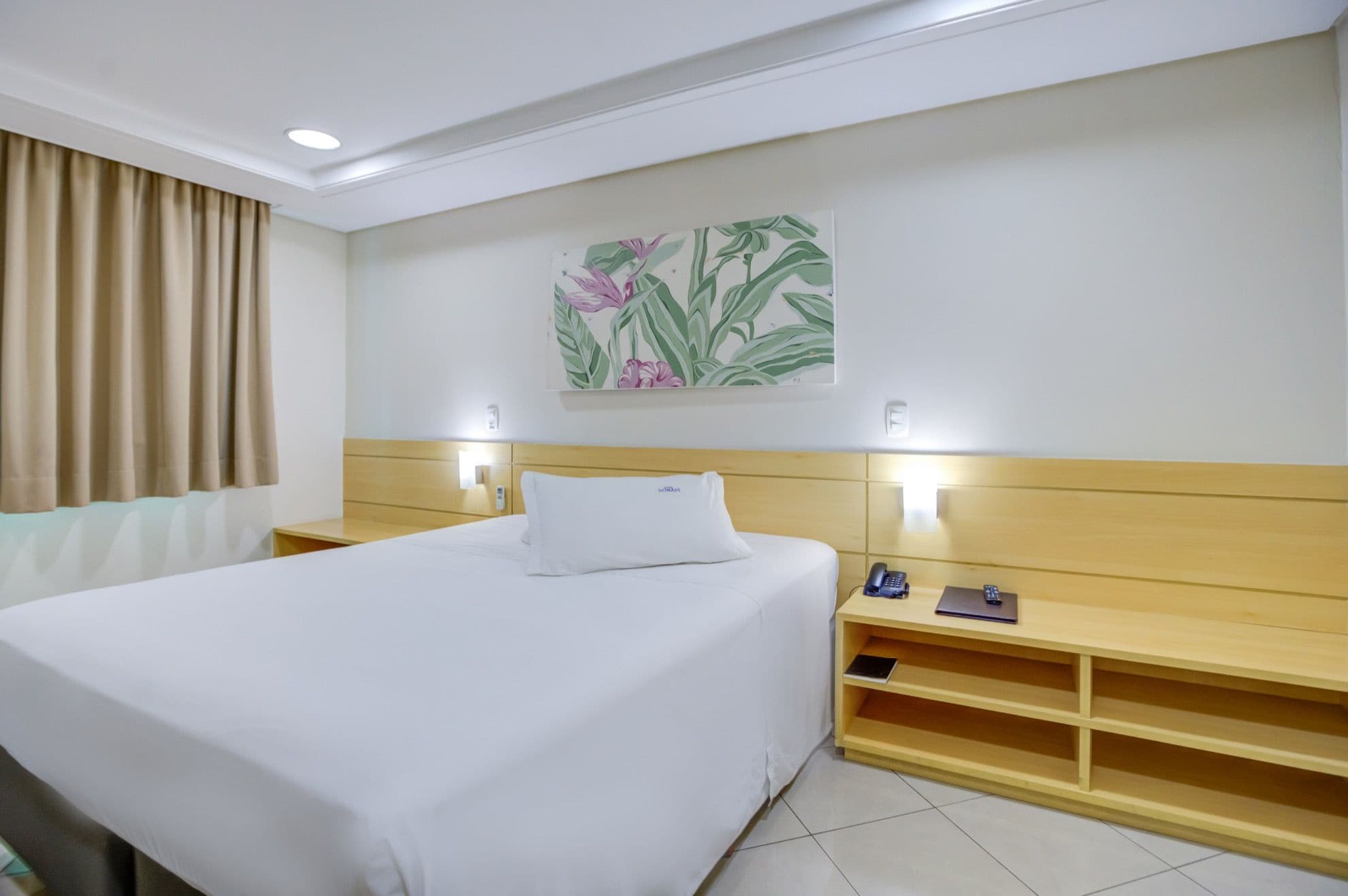 Habitació d'hotel amb un llit gran, taules de nit, un telèfon fixat a la paret i una petita estanteria amb un quadern i una ploma.