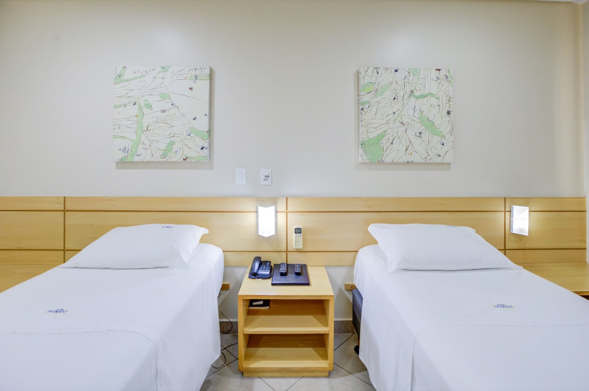 A hotel room with two single beds, a wooden nightstand with a phone and remote controls, and two abstract wall art pieces.