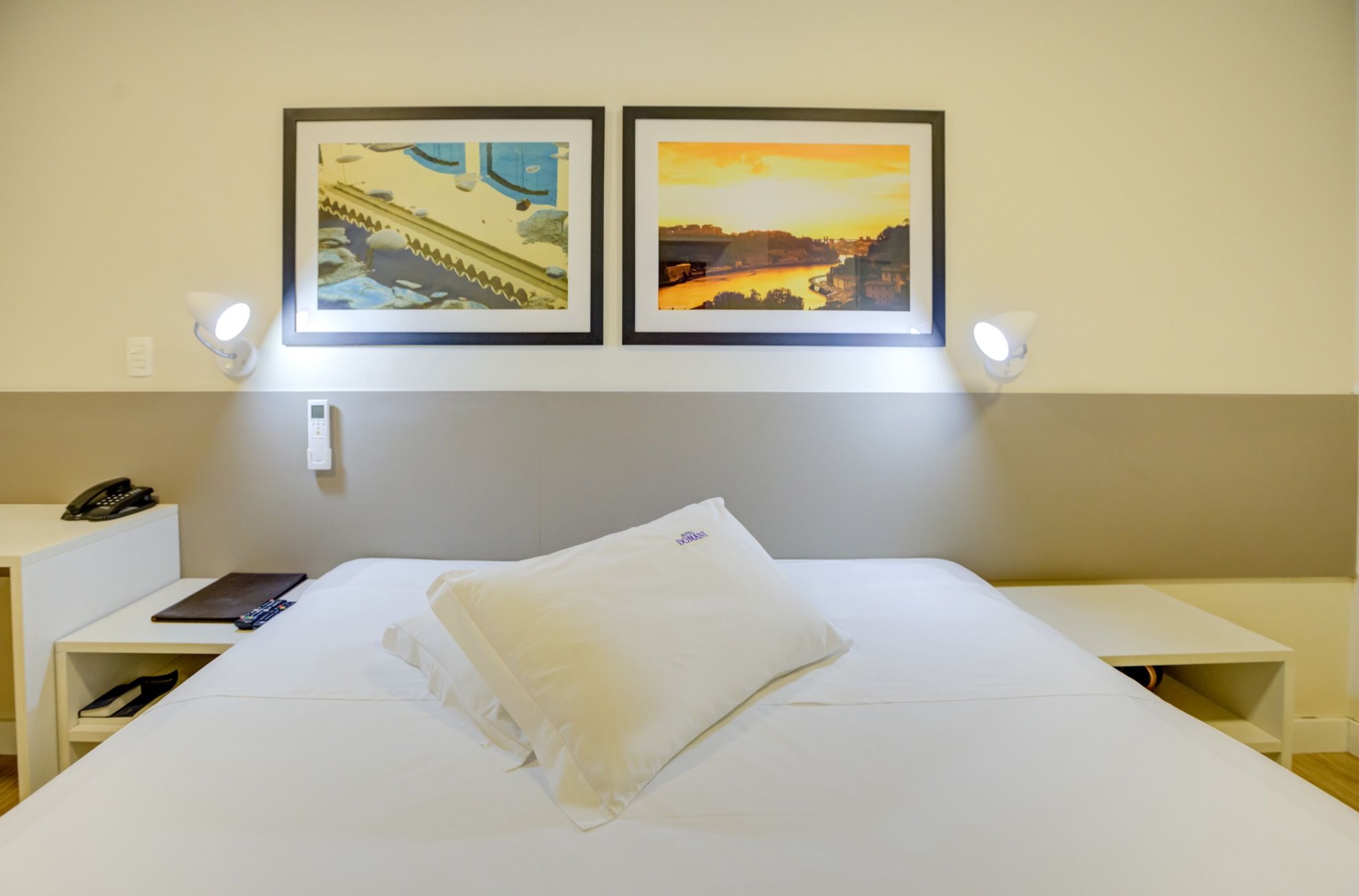 A hotel room with a bed, a bedside table with a phone, and two framed pictures on the wall.