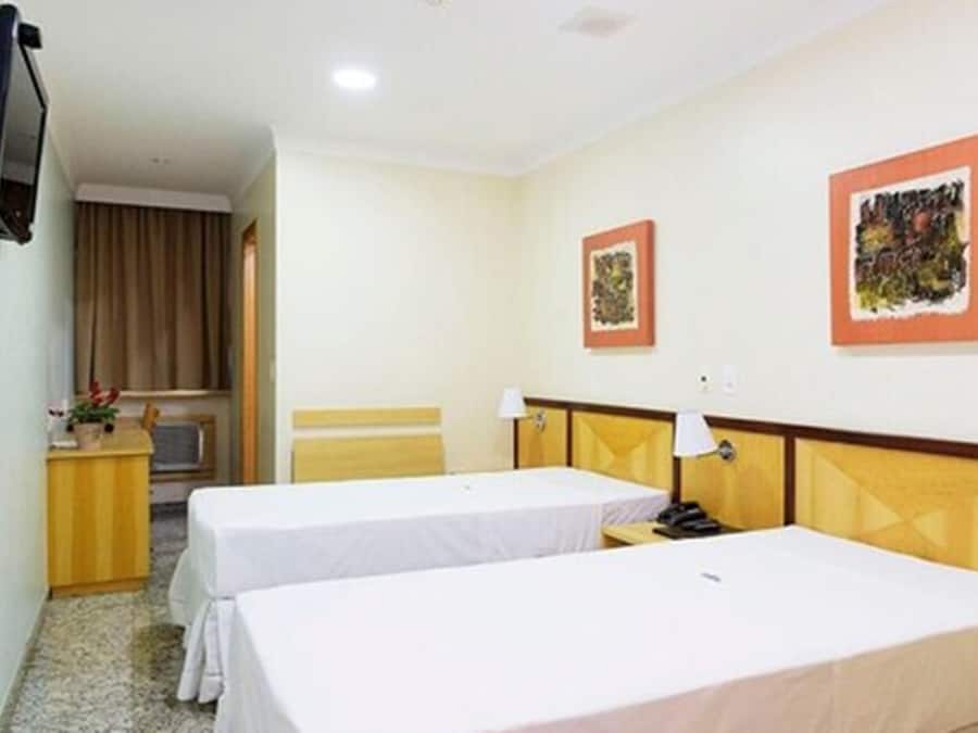A hotel room with two single beds, a desk with a chair, a television mounted on the wall, and two framed pictures on the wall.