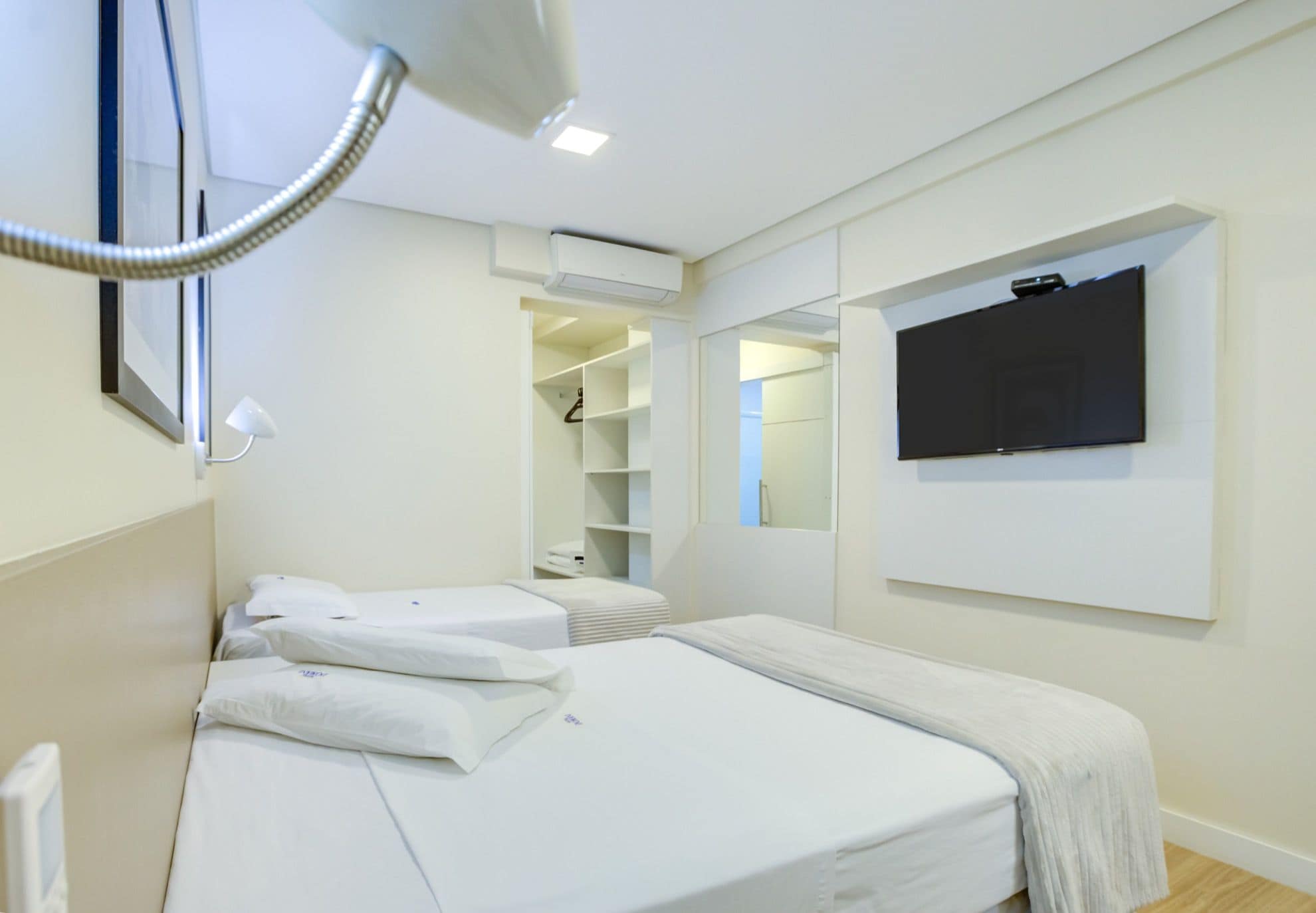 Una habitación de hotel con cama, televisión montada en la pared, vestidor y aire acondicionado instalado en el techo.