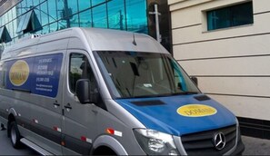 A silver van with a blue front, parked in front of a building with large windows. - Hotel Domani (Guarulhos)