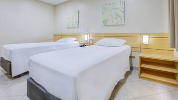 A hotel room with two single beds, a wooden headboard, a small wooden shelf, and two wall-mounted light fixtures.