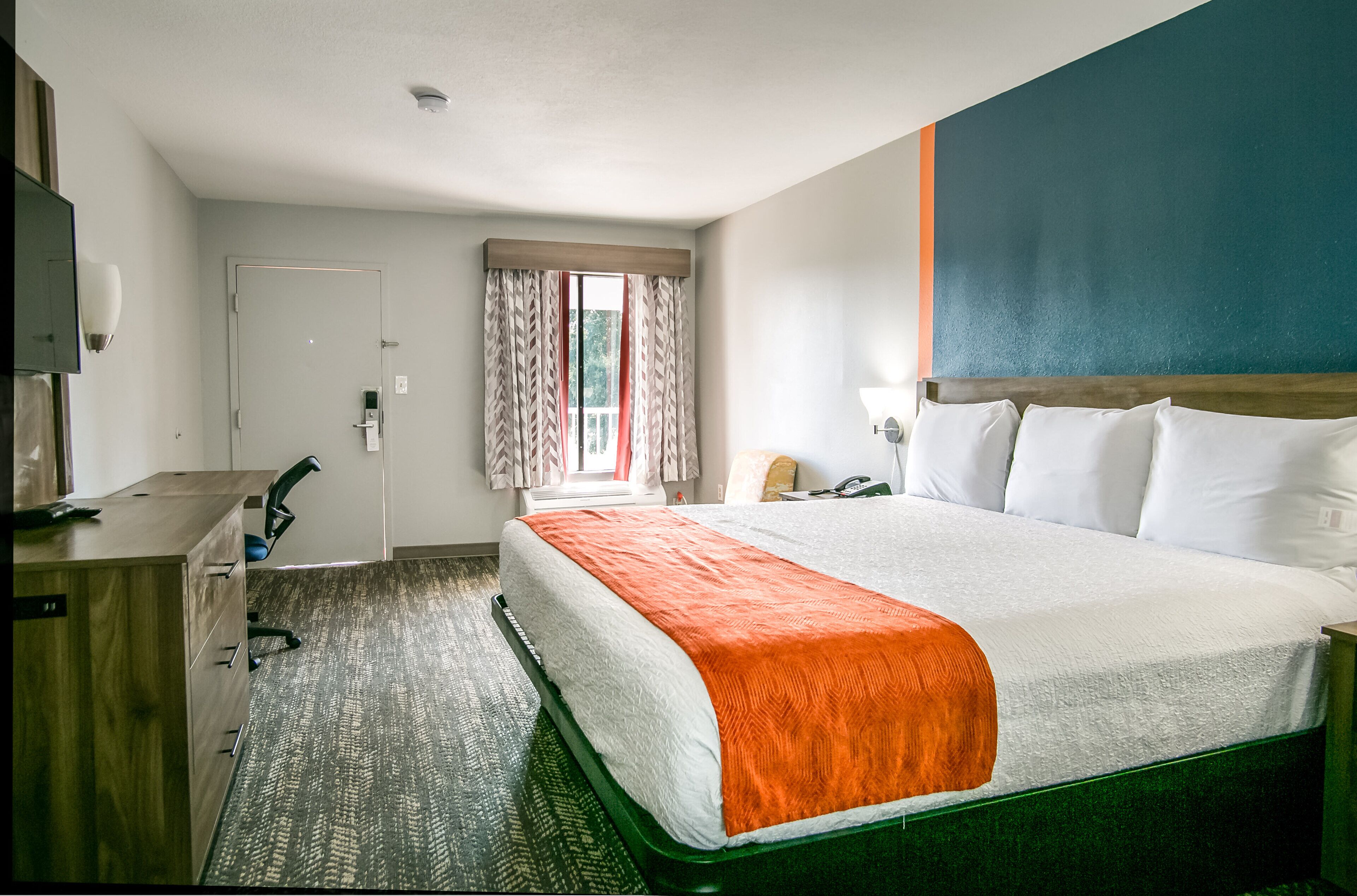 A hotel room with a bed, a desk, a chair, a window with curtains, and a wall with a painting.