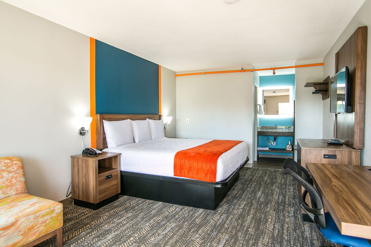 A hotel room with a bed, a desk, a chair, a TV, and a bathroom visible through an open door.