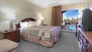 Ein Hotelzimmer mit einem Bett, einem Fernseher, einem Schreibtisch mit Lampe, einem Sessel und Blick auf einen Pool und Palmen.