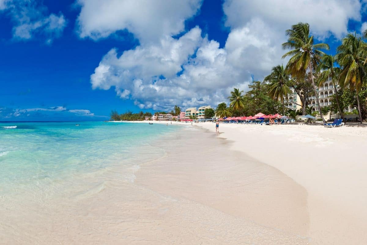 A beach with clear turquoise water, white sand, and palm trees. There are buildings with balconies and umbrellas in the background.
