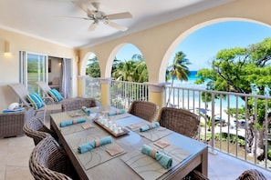 A dining area with a view of the beach, featuring wicker furniture, a ceiling fan, and a table set for four. - Sapphire Beach Condominiums by Blue Sky Luxury (St. Lawrence Gap)