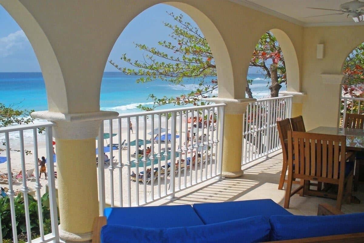 A balcony with a view of the beach and ocean, featuring a table and chairs, and a view of the sea through arches.