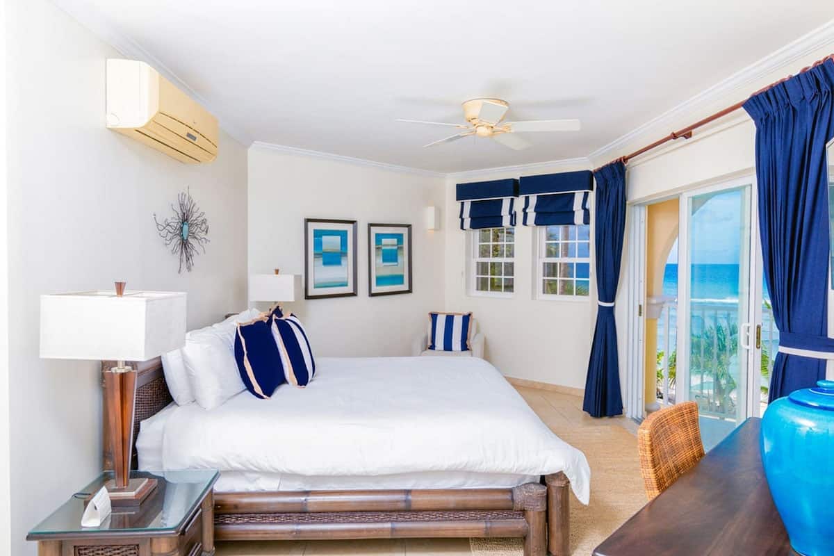 A hotel room with a bed, a nightstand, a lamp, a ceiling fan, a window with curtains, and a view of the ocean.