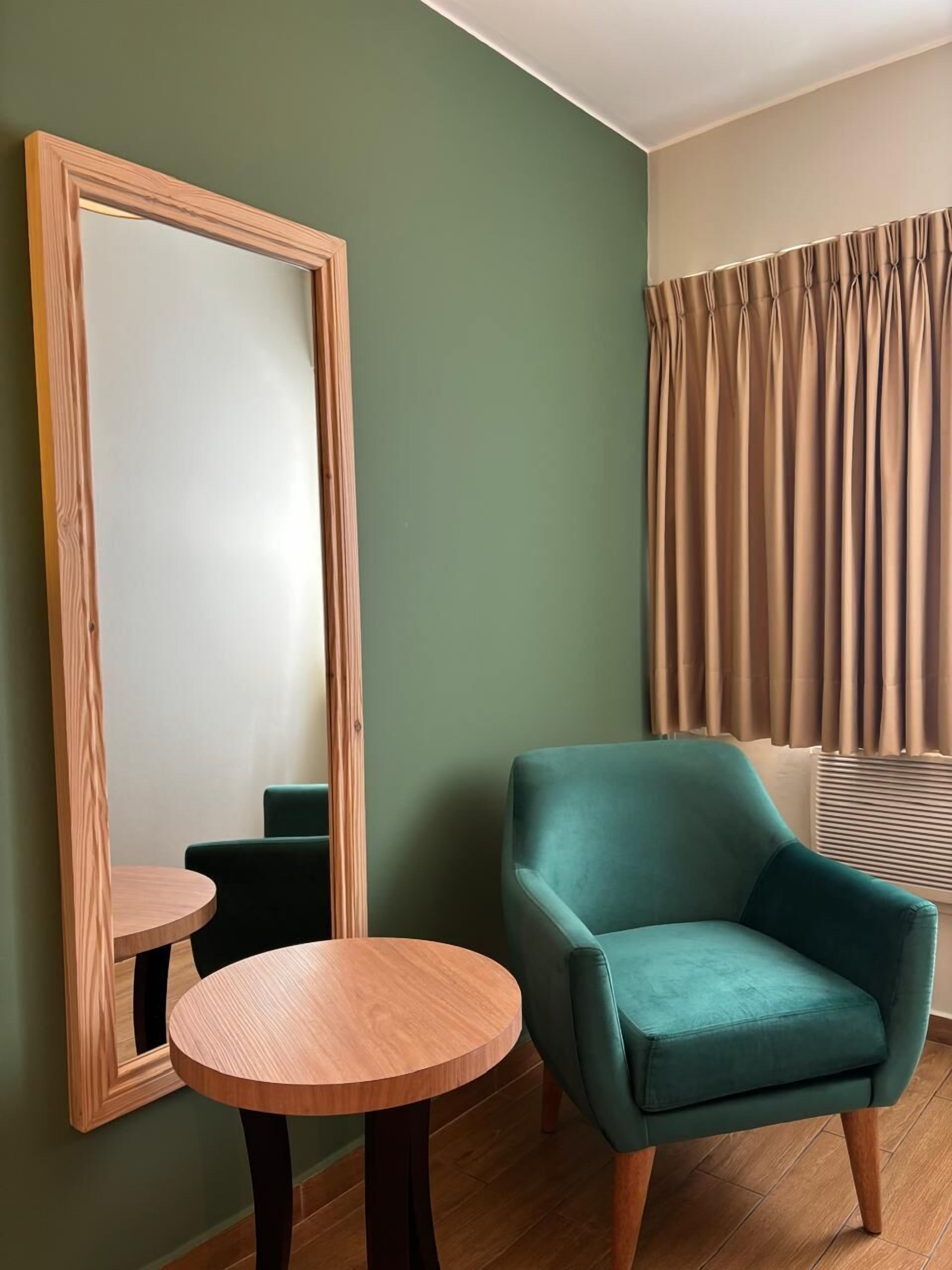 A room with a large mirror, a teal armchair, a small wooden table, and a window with curtains.