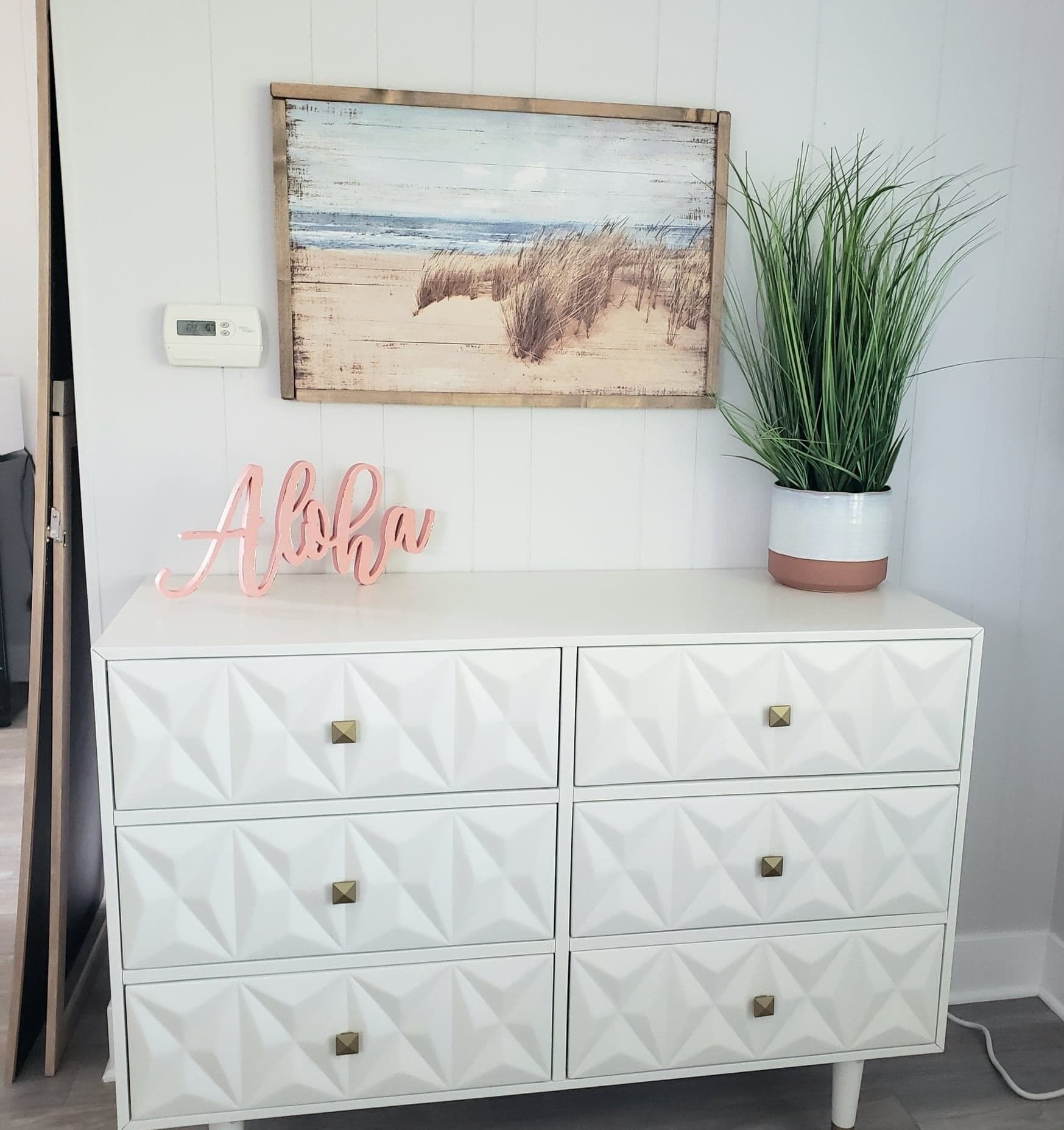 A white dresser with geometric patterns and gold handles, a pink 'Absolu' sign, a beach painting, and a potted plant.