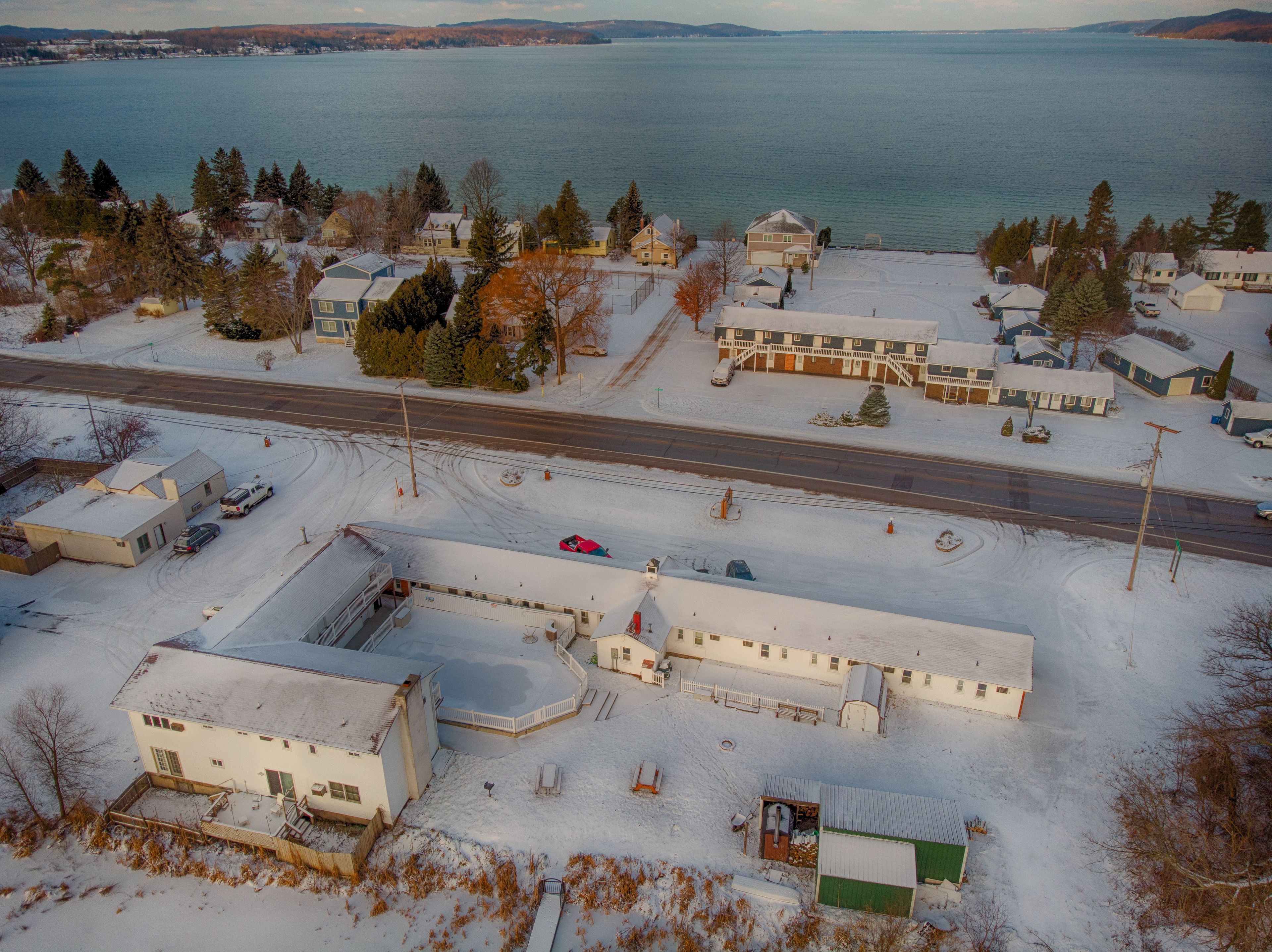 A snow-covered residential area with houses, a road, and a body of water in the background.