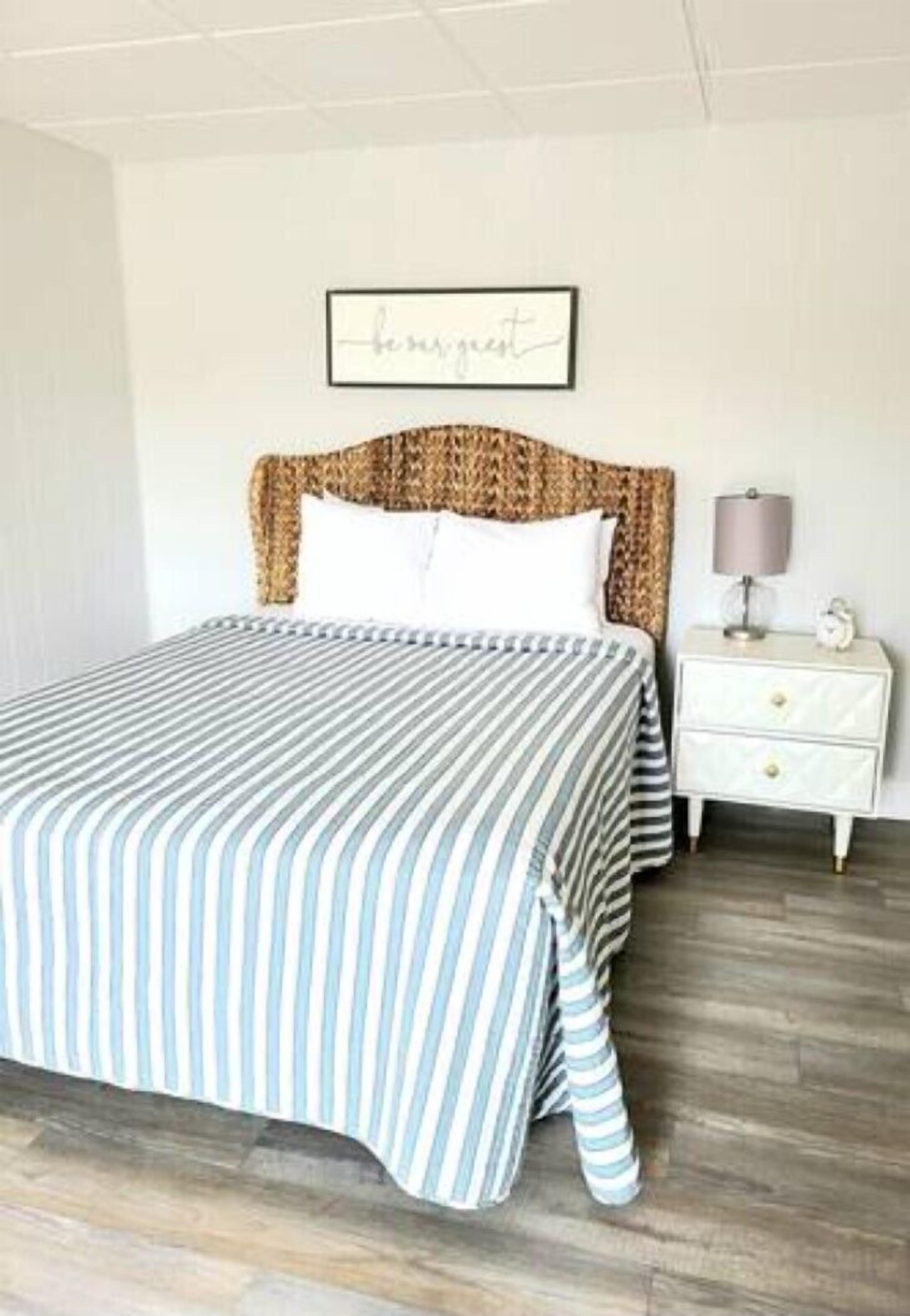 Un lit bien fait, avec une literie à rayures, une tête de lit ornée d’un motif tissé, une table de chevet avec une lampe et un cadre décoratif accroché au mur.