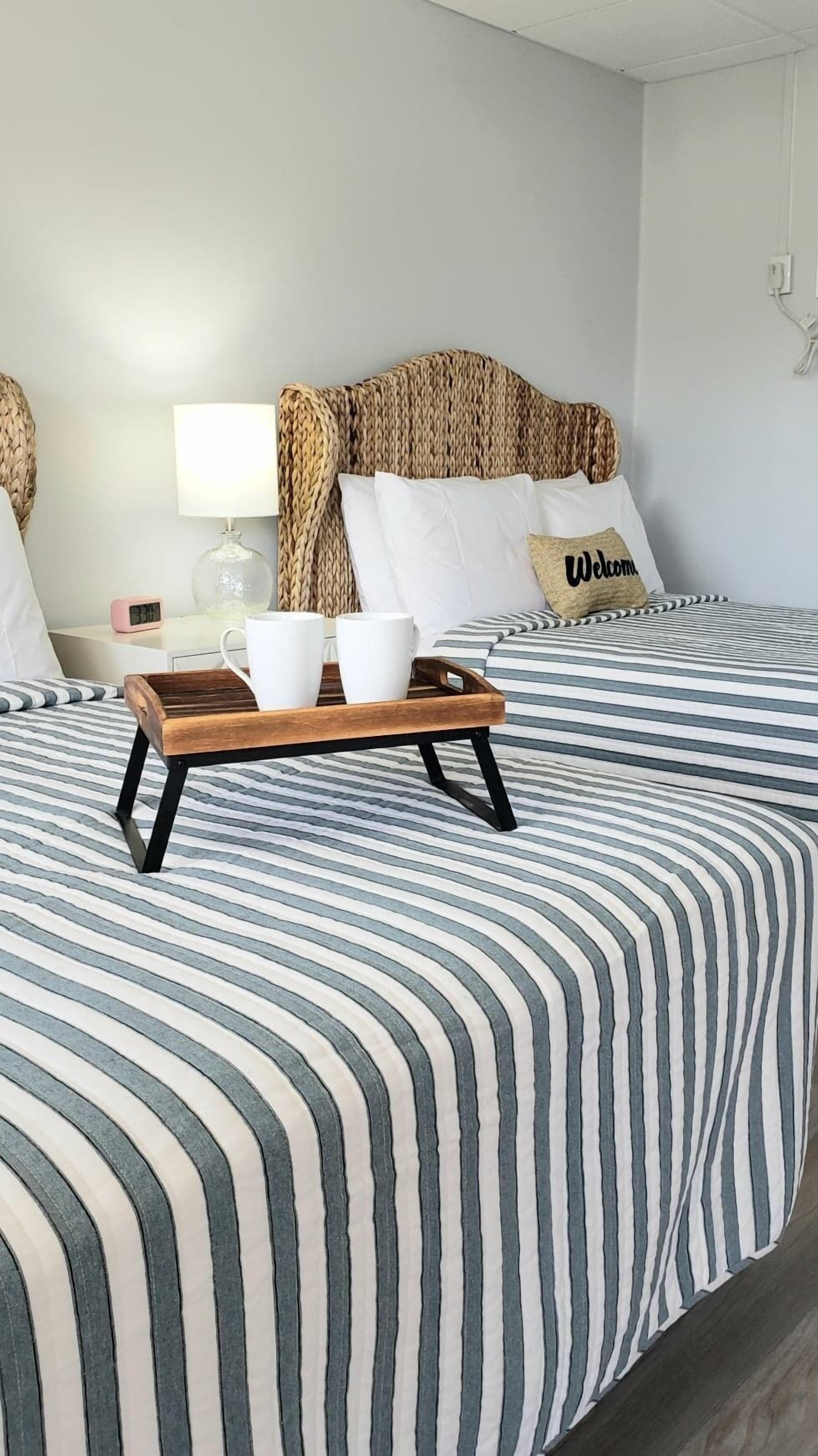 A hotel room with a bed featuring striped bedding, a wooden tray with two white mugs, a lamp, and a pillow with 'Welcome' text.