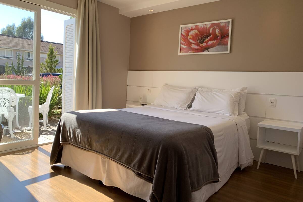 A bedroom with a large bed, a bedside table, a nightstand, and a window with a view of greenery and buildings.