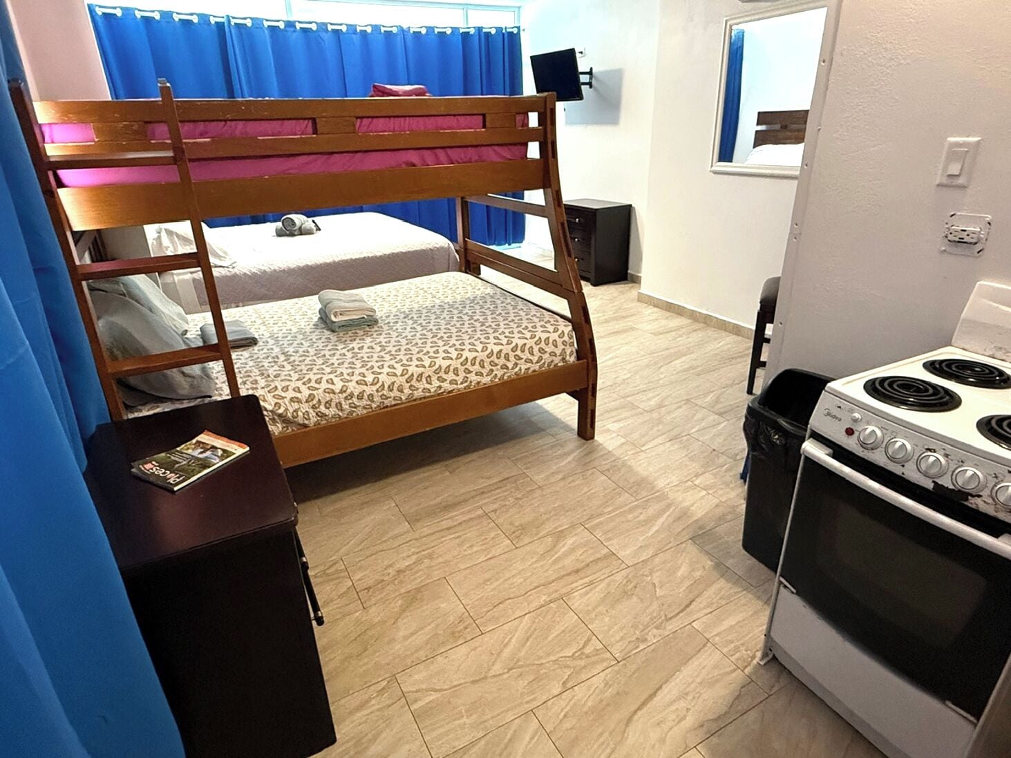 Uma cama beliche com colchão estampado, uma pequena mesa de madeira com uma revista, um fogão e uma cortina azul.