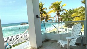 A balcony with a view of the ocean and palm trees, white chairs, and a tiled floor.