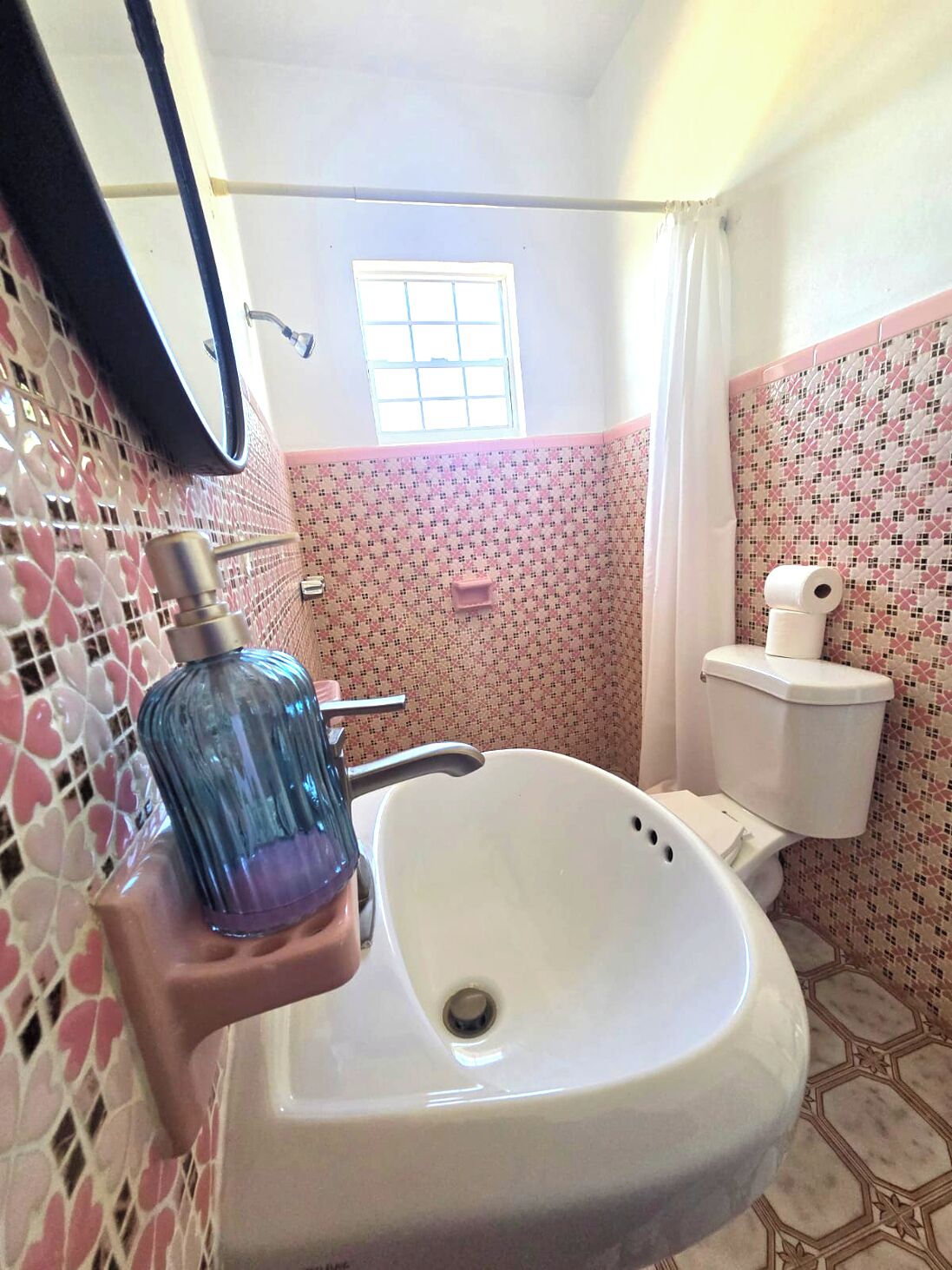 A bathroom with a white sink, a mirror, a window, and pink patterned walls.