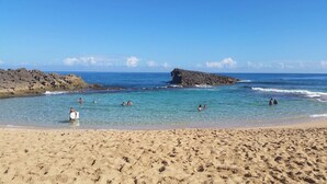 A sandy beach with clear turquoise water, rocky formations, and people swimming and relaxing.
