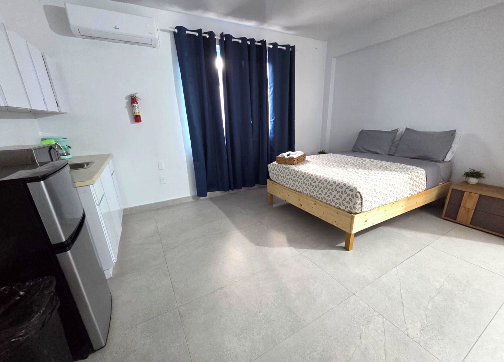 Une chambre compacte comprenant un lit, une petite table de chevet, une commode, une fenêtre avec des rideaux et un coin cuisine équipé d’un réfrigérateur et d’une cuisinière.