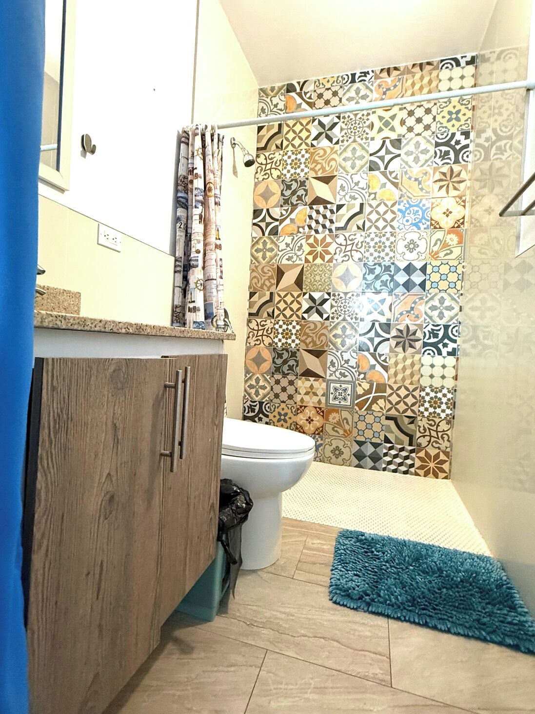 A bathroom with a toilet, a shower with a patterned tile wall, a blue rug, and a wooden cabinet.