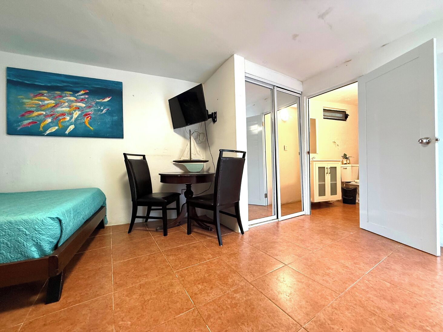A small room with a bed, a table with chairs, a painting, and a sliding door leading to a bathroom.