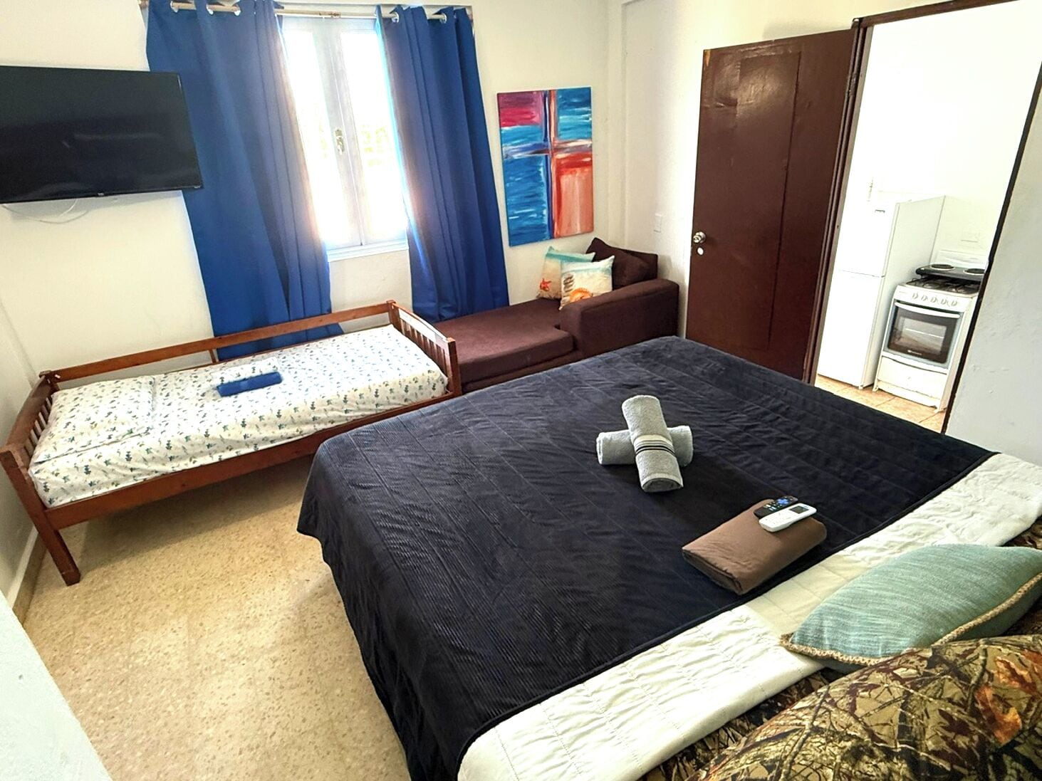 Un dormitorio con cama, un banco de madera, televisor, un cuadro y una kitchenette.