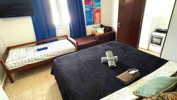 Un dormitorio con cama, un banco de madera, televisor, un cuadro y una kitchenette.