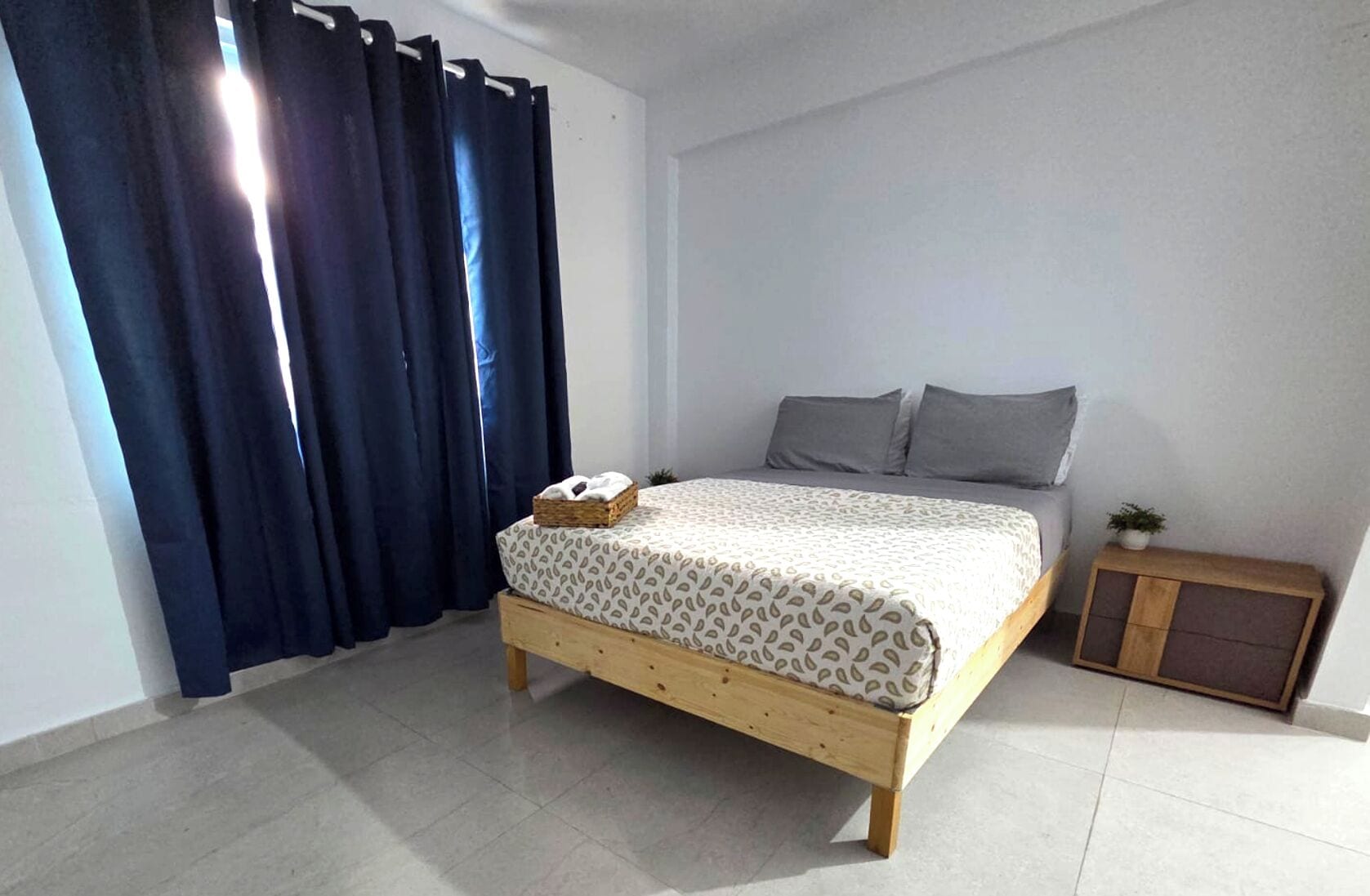 A bedroom with a bed, curtains, a small wooden cabinet, and a tiled floor.