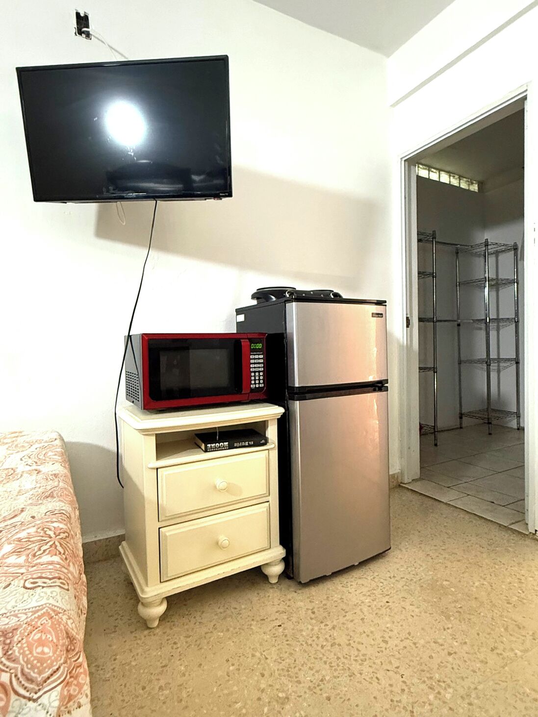 A compact hotel room with a kitchenette, a flat-screen TV mounted on the wall, and a bed with patterned bedding.