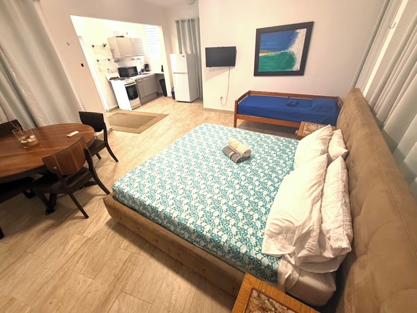 Una habitación con cama, una mesa de madera, una silla, una kitchenette pequeña y un cuadro en la pared.