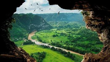 A view through a cave opening, revealing a river winding through a lush green valley with mountains in the background.
