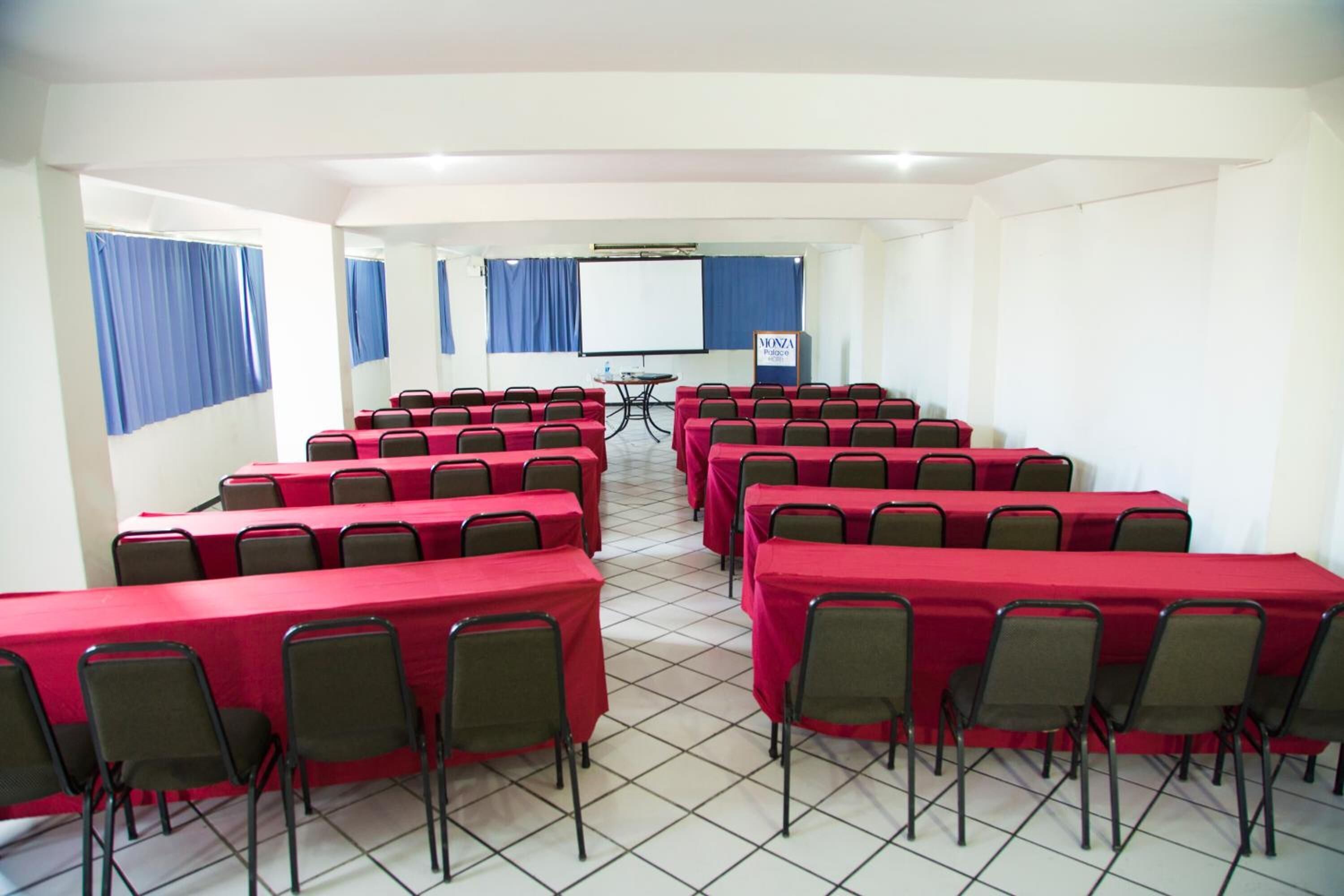Un salón de conferencias con varias mesas y sillas, una pantalla de proyector y una pizarra.