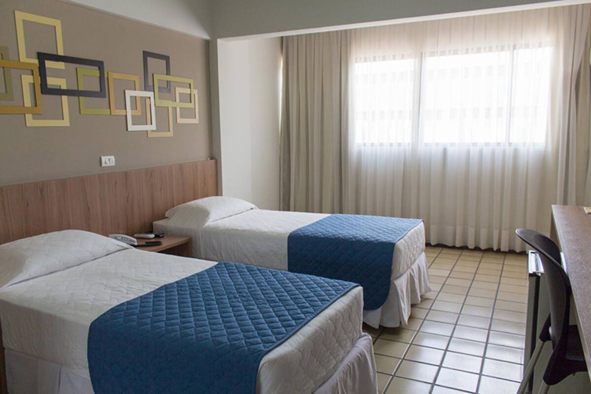 A hotel room with two beds, a wooden headboard, a desk, and a window with curtains.