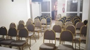 Un salón de conferencias con filas de sillas y mesas, un extintor en la pared y una ventana al fondo.