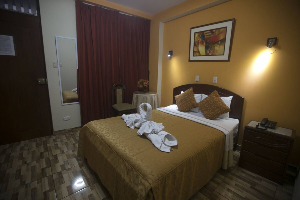 Quarto de hotel com cama, mesa de cabeceira, cadeira, pequena mesa com um vaso e um quadro na parede.